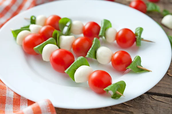 Caprese traditional mediterranean salad skewers with mozzarella basil and tomatoes on white dish. Vintage wooden table background. — Stock Fotó