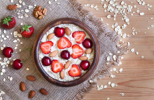 Delicious natural breakfast oatmeal porridge with strawberry, nuts and cherry in wooden bowl on vintage table background. — Stock Photo, Image