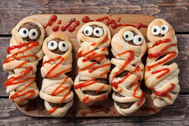 Halloween homemade food sausage meatball mummies wrapped in dough, baked and covered with fake blood sauce decoration for holiday celebration party on vintage wooden background.