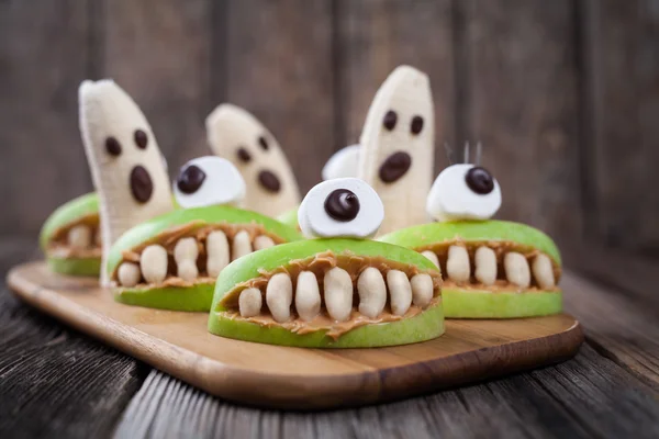 Scary edible halloween treat apple cyclop mouth with peanut butter teeth and banana ghosts chocolate face. Healthy natural vegetarian dessert recipe. — Stock Photo, Image