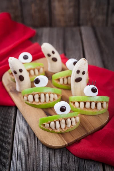 Scary Halloween alimentos comestibles monstruos celebración saludable fiesta decoración receta. Loco cíclope boca de manzana con dientes y fantasmas de plátano, tela roja sobre fondo de madera vintage . — Foto de Stock