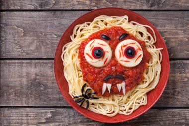 Halloween party decoration food. Spaghetti monster face with big eyeballs, fangs, spider and moustaches in red dish on vintage wooden table background clipart