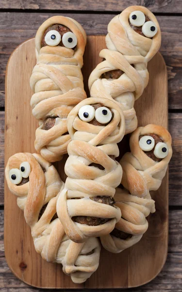 Scary halloween food sausage meatball mummies in dough baked for october celebration party decorated with funny eyes. Vintage wooden background. — Stock Photo, Image