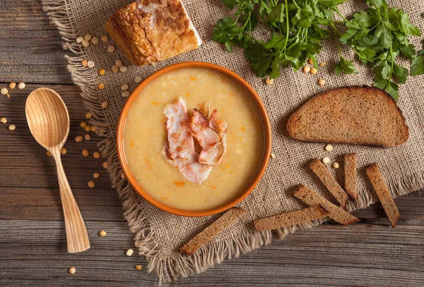 Homemade traditional split pea soup recipe food composition with bacon, croutons, greens, ham and wooden spoon on vintage table background. — Stock Photo, Image