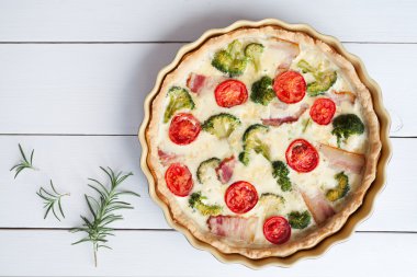 Quiche lorraine tart pie traditional homemade french food preparation recipe with cheese tomatoes broccoli and bacon in baking dish on white wooden table background.