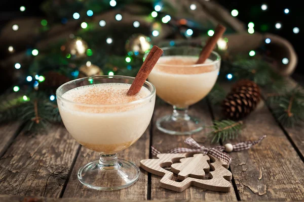 Eggnog traditional xmas homemade winter egg, milk, rum, vanilla alcohol liqueur preparation recipe in two glass cups with cinnamon sticks on wooden vintage table. Blue and green bokeh background — Stock fotografie