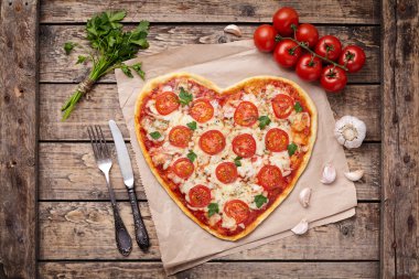 Heart shaped pizza margherita love concept for Valentines Day with mozzarella, tomatoes, parsley and garlic on vintage wooden table background.
