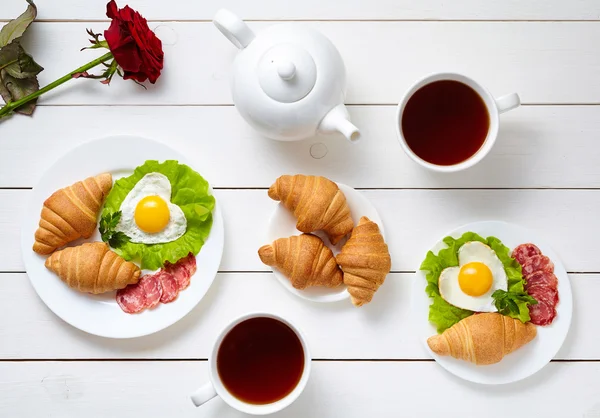 Romantic breakfast for Valentines Day with heart shaped eggs, salad, croissants, rose flower and tea on white wooden table background. Food concept of love. — Zdjęcie stockowe