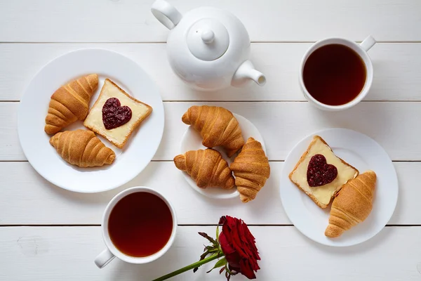 Micul dejun romantic pentru doi cu pâine prăjită, gem în formă de inimă, croissante, trandafir și ceai pe fundal de masă albă din lemn . — Fotografie, imagine de stoc