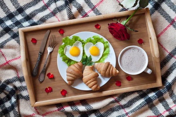 Romantic breakfast in bed with heart-shaped eggs, salad, croissants, coffee, rose flower and petals on wooden tray. — Stock Photo, Image