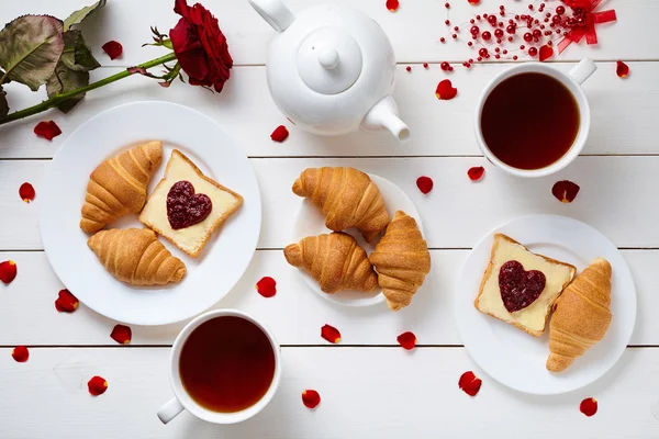 Romantic breakfast for Valentines day with toasts, heart shaped jam, croissants, rose petals and tea on white wooden table background. — Stock Fotó