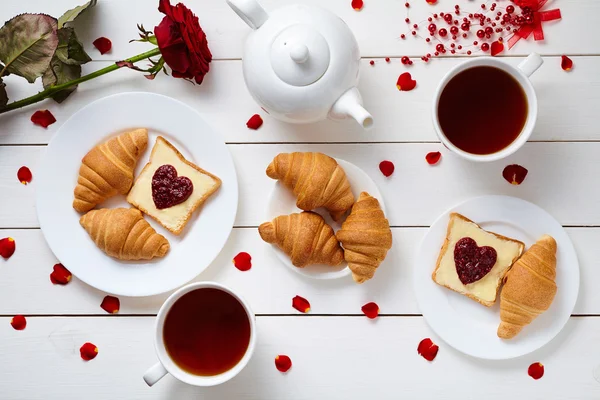 Romantic breakfast for Valentines day with toasts, heart shaped jam, croissants, rose petals and tea on white wooden table background. — Stock Photo, Image