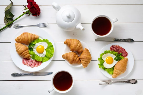 Romantisches Frühstück für zwei Personen mit herzförmigen Eiern, Salat, Croissants, Rosenblüten und schwarzem Tee auf weißem Holztischhintergrund. Ernährungskonzept der Liebe. — Stockfoto