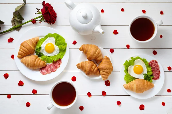 Breakfast for Valentines day with heart shaped eggs, salad, croissants, rose petals and tea on white wooden table background. — 스톡 사진