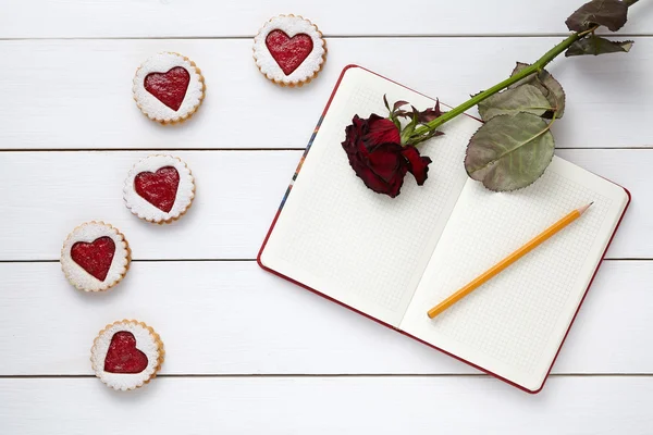 Biscotti a forma di cuore con quaderno vuoto, matita e fiore di rosa su sfondo bianco in legno per la celebrazione di San Valentino . — Foto Stock