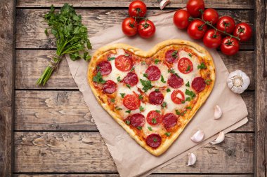 Heart shaped pizza with pepperoni, tomatoes, mozzarella, garlic and parsley composition on vintage wooden table background.