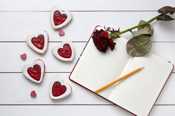 Biscotti a forma di cuore con quaderno vuoto, matita e regalo di rosa rossa su sfondo bianco in legno . — Foto Stock