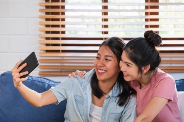 Mutlu Asyalı lezbiyen çift, arkadaşlarıyla evdeki oturma odasındaki kanepede görüntülü konuşma yapıyor.