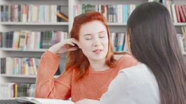 Üniversite kütüphanesinde okurken arkadaşıyla sohbet eden genç bir kadın.