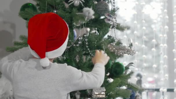 Chico encantador en el sombrero de Papá Noel decorando el árbol de Navidad — Vídeo de stock