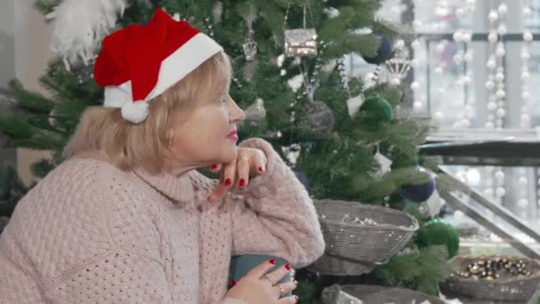 Mujer mayor sonriendo a la cámara mientras está sentada cerca del árbol de Navidad en casa — Vídeos de Stock