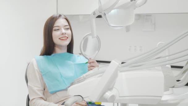 Весела молода жінка посміхається до камери, сидячи в стоматологічному кріслі — стокове відео