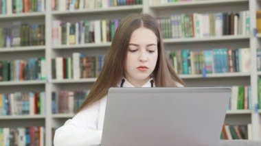Genç kız kütüphanede dizüstü bilgisayarla çalışırken dikkatle başka tarafa bakıyor.