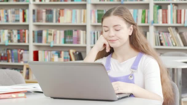 Fáradt tini lány dolgozik megbízást a laptopján a könyvtárban — Stock videók