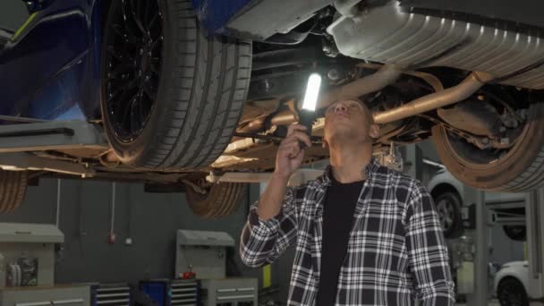 Hombre guapo examinando coche levantado con una lámpara en el taller — Vídeos de Stock