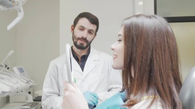 Diş tedavisi sonrası aynaya bakan bir dişçi hastayla konuşuyor.