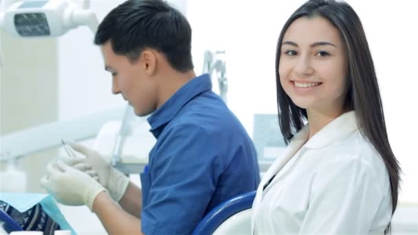 Девушка-ассистентка сидит рядом со стоматологом и улыбается зубам прямо перед камерой — стоковое видео
