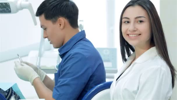 Asistente de chica sentada junto a un dentista y sonriendo directamente a la cámara, luego pulgar hacia arriba — Vídeo de stock