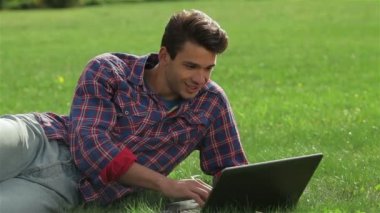 Parkta dizüstü bilgisayarla çimenlerin üzerinde yatan adam