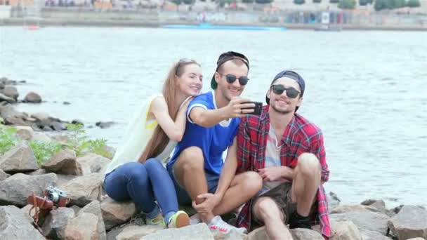 Drie vrienden zitten en lachen aan de rivier en foto's van elkaar — Stockvideo