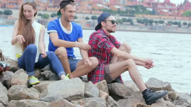 Трое друзей сидят, смеются и бросают камни в реку — стоковое видео
