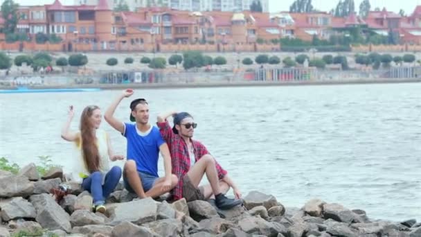 Três amigos sentados e rindo e jogando pedras no rio — Vídeo de Stock