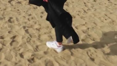 Kadın bacakları kumlu sahil boyunca yürür, beyaz spor ayakkabılarla, ellerinde deri bir ceket
