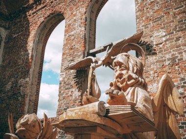 Kırmızı tuğlalı bir bina, harabeler, elinde tüy olan ahşap bir melek ve kaidede bir kitap, ahşap bir leylek ve ahşap bir gülün arkasında, büyük boş pencerelerin arasından bulutlu mavi bir gökyüzü çıkıyor.