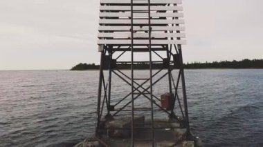 Küçük bir demir deniz feneri ve beyaz tahtalar, gölün etrafındaki kayalık yüzeyde, merdivenler yukarı, dron düzgünce yükseliyor, gökyüzü bulutlu, beyaz
