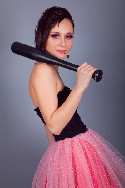 Elegantní brunetka s náušnice v růžové a černé šaty s baseballovou pálkou v ruce na šedém pozadí — Stock fotografie