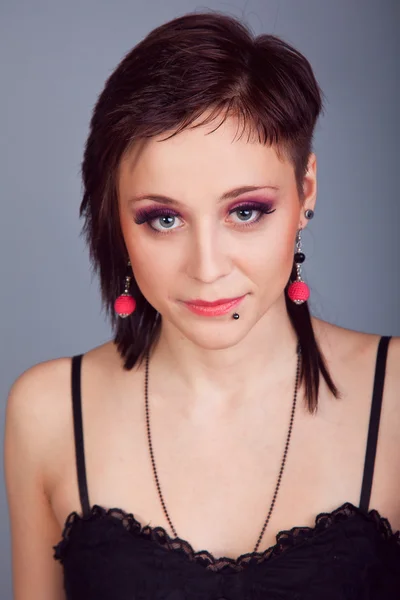 Portrait of a beautiful brunette girl with short hair with red earrings in a black dress on a gray background — Stock Photo, Image