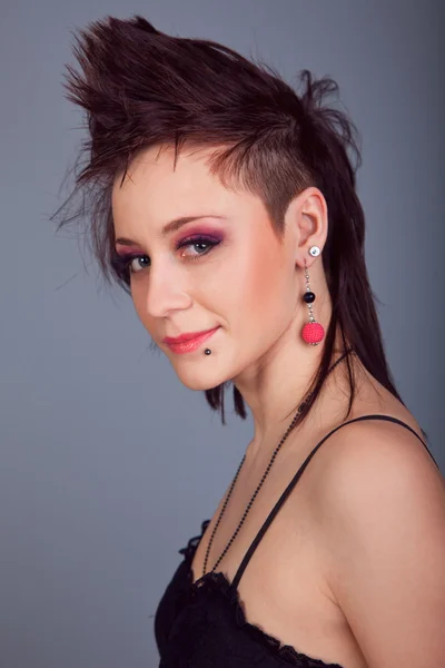 Portrait d'une belle fille brune avec mohawk en boucles d'oreilles rouges dans une robe noire sur un fond gris — Photo