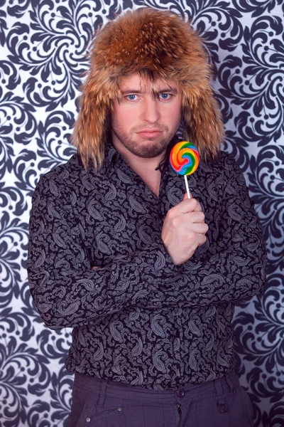 Portrait of serious business man in black shirt with patterns in a fur hat with lollipop — Stock Photo, Image