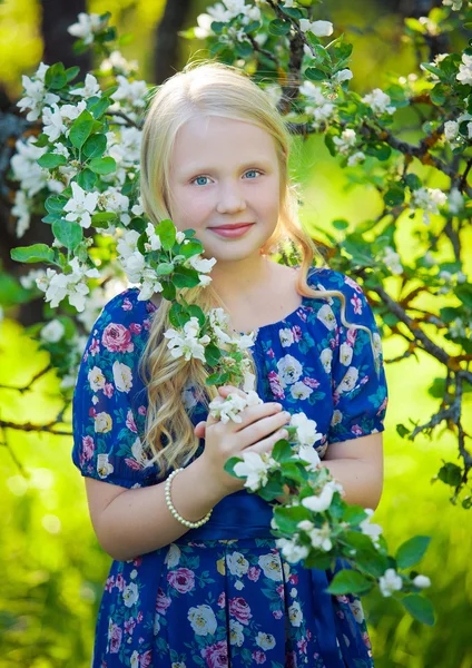 Adorable toddler girl with curly hair and flower crown wearing a magic fairy costume with a blue dress and angel wings playing in a sunny blooming fruit garden with cherry blossom and apple trees.