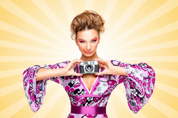 Retro smile. Retro photo of a smiley fashionable hippie homemaker, holding an old vintage photo camera with two hands and showing emotions on colorful abstract cartoon style background. — Stock Photo, Image