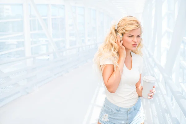 Morgenspaziergänge. schöne junge Frau mit Musikkopfhörern, die mit einer Kaffeetasse zum Mitnehmen auf der Brücke steht und vor urbanem Hintergrund posiert. — Stockfoto