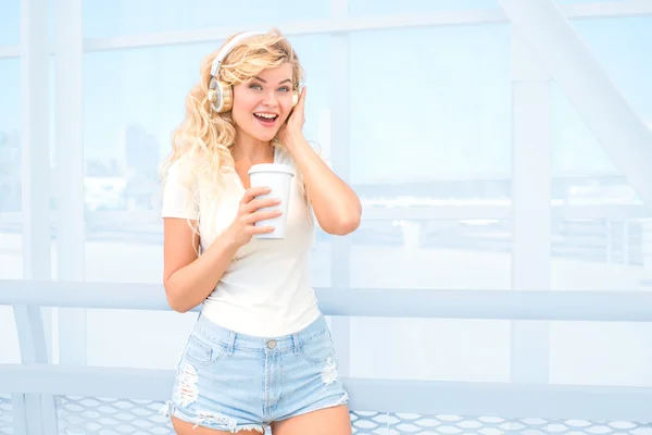 Paseos matutinos. Hermosa joven con auriculares musicales, de pie en el puente con una taza de café para llevar y posando sobre fondo urbano . — Foto de Stock