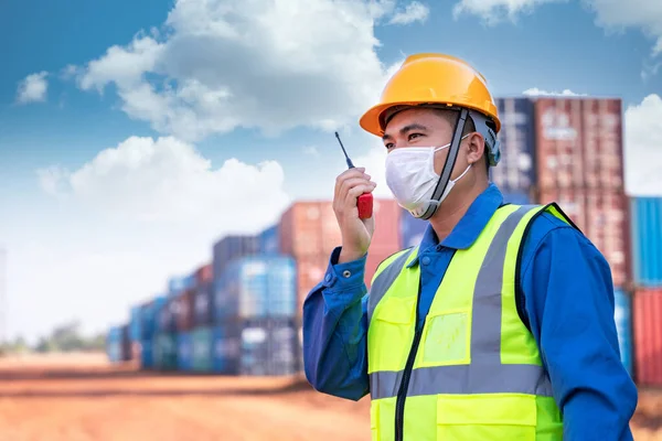 Asian foreman worker wear a mask working use a long distance radio control container cargo harbor to loading containers prevent the spread of the coronavirus. Logistics import export shipping concept.