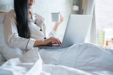 Dizüstü bilgisayar kullanan ve çalışan, yatak odasındaki beyaz bir yatakta oturup kahve içen bir kadının yakın çekimi..
