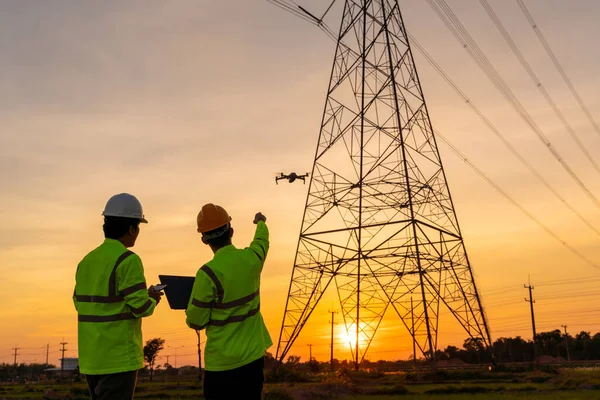 エンジニアのチームの作業場所ヘルプ技術者は 高圧送電塔 発電所の労働者ではなく 飛行中の機器を検査するドローンを使用するのに役立ちます — ストック写真